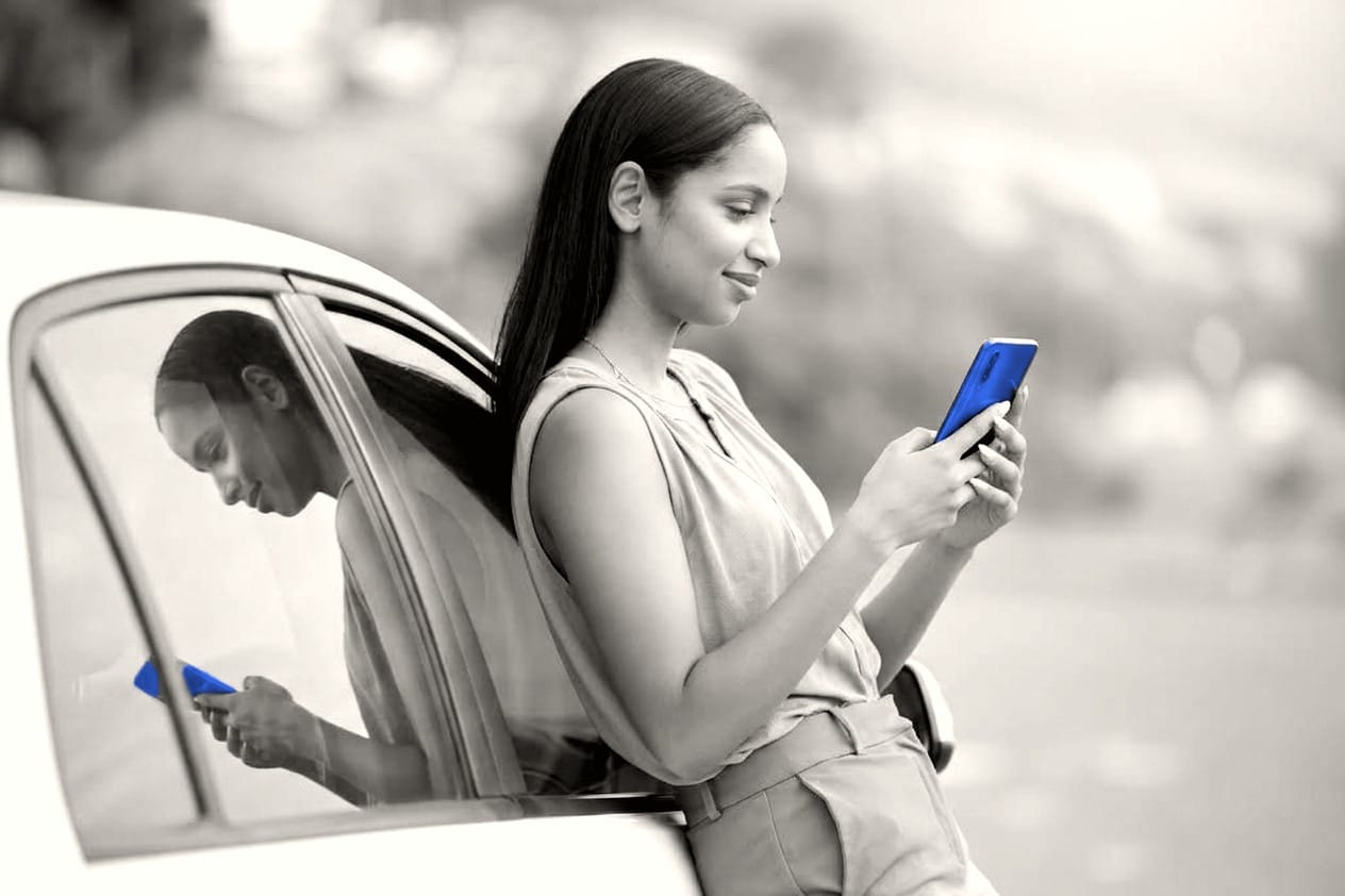 Kundin lehnt an ihrem Auto und erledigt die Zulassung am Smartphone